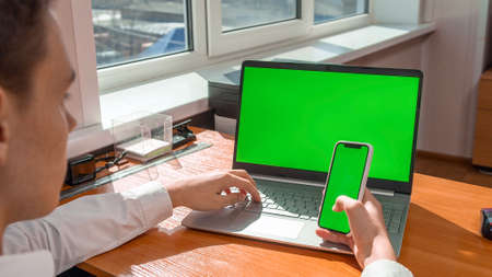 A young man works in the office behind a laptop with a green screen, in his hands holds a phone with...