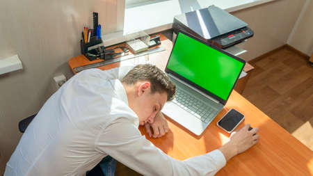 A tired young Caucasian businessman fell asleep at his desk in the office in front of a computer with a green screen on the monitor. High quality photoの写真素材
