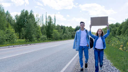A young couple walks by the side of the road with a cardboard box in their hands with a place for text, hitchhiking, traveling, adventures. High quality photoの写真素材