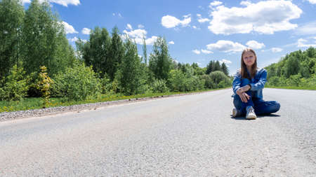 A smiling young girl, a teenager, sits in the middle of the road in the summer outside the city. The concept of tourism and travel advertising. High quality photoの写真素材