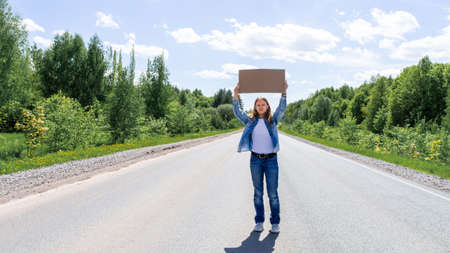 A young girl, a teenager with a backpack and a cardboard box for text, is standing in the middle of the road. The concept of travel, a passing car, vacation. Place for text. High quality photoの写真素材