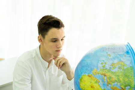 A thoughtful student in a white shirt is sitting at a table in front of a globe. The concept of geography lesson, travel. High quality photoの写真素材