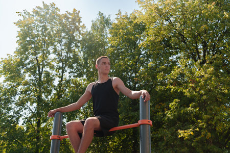 A young guy sits on a horizontal bar with his legs dangling in a city park on a summer day.