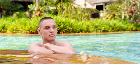 A young guy at the edge of the pool enjoys the cool blue water of the hot summer. In the background there is a green planting of flowers.の写真素材