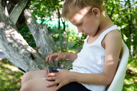 blonde-haired child engrossed in the use of a handheld device, his hunched posture potentially straining his developing spine as he remains sedentaryの写真素材