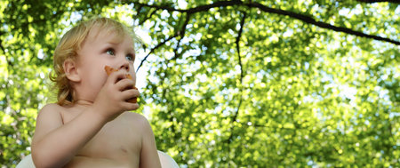 Banner, curious baby with golden curls is eating a tasty cookie, his face looking curiously into the distance, copy spaceの写真素材