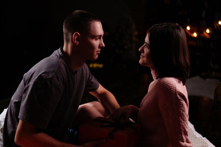 Young couple in love, guy giving a gift to girl in semi-darkened living room, concept new year,...