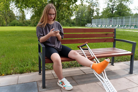 Young female with a casted leg relaxing on a bench in a lush summer park, reading content on her...