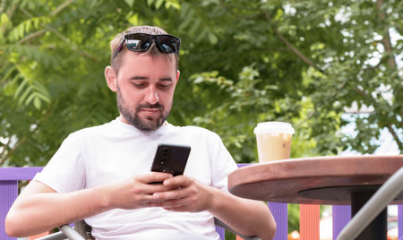 Bearded man wearing sunglasses and a white t shirt, sipping iced coffee at an outdoor cafe, using a...