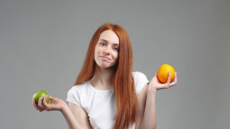 Portrait of a young girl standing on a gray background wondered what fruit is healthier Apple or orange.の写真素材