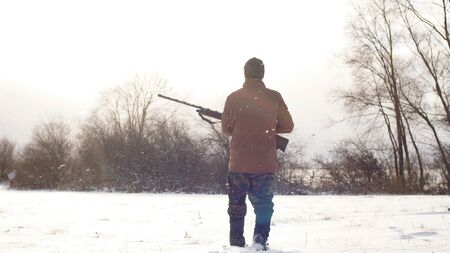 man dressed in warm clothes is walking across the field with a hunting rifle.