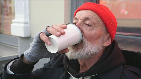 Bearded old man, homeless man drinking a hot drink to keep warm. A tired homeless man drinks tea sitting on a cardboard on the street.の写真素材