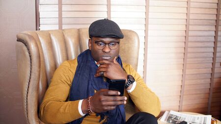 young African-American man in glasses and a cap is sitting in a chair of an expensive cafe,...