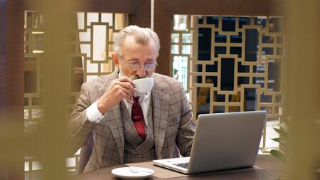 well-Groomed, elegant, stylish, fashionable and bearded old publisher, writer, entrepreneur, Director, dressed in an elegant jacket, sitting in a restaurant at a laptop and drinking tea and coffee.の写真素材