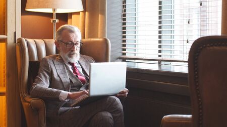 Elegant, charismatic old man, gray-haired pensioner, millionaire, elderly man sitting at a laptop and drinking coffee, tea.の写真素材