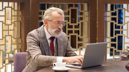 Fashionable, stylish old man, retired, gray-haired man with a beard sitting at a table and working...