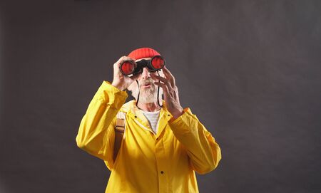 Happy curious man, the traveler looks into the distance, uses binoculars, wears a red hat and yellow Cape, isolated background.の写真素材
