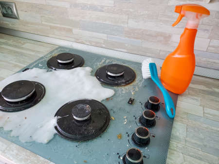 Photos of a dirty modern gas stove covered in foam, with detergent and a brush for cleaning the stove in the background.の写真素材