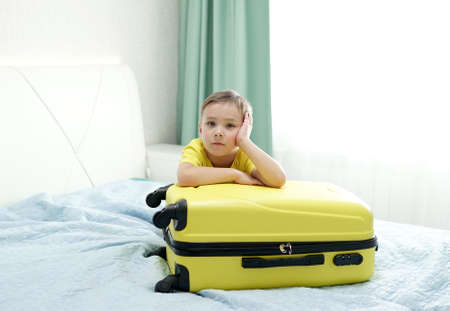 Child with blond hair in a yellow t-shirt is waiting for a vacation and dreams of the sea, putting his hands on a suitcase.の写真素材