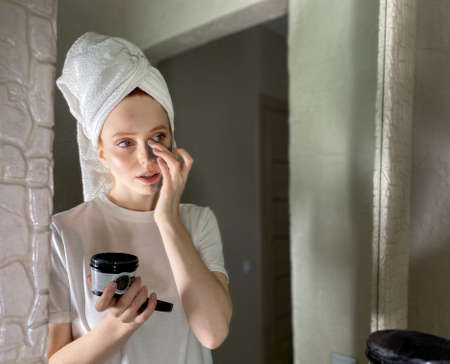close-up of a young woman with a towel on her head, applies a cleansing mask on her face made of gray clay in front of a mirror.の写真素材