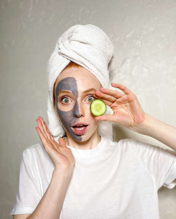 young woman has applied a gray clay mask to half of her face, posing for the camera holding a slice of fresh cucumber in her hand.の写真素材