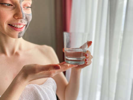 pretty young woman with a mask on her face holding a glass of clean water in her hands, close-up. beauty treatments at home.の写真素材