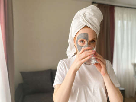 face Care at home concept. a young woman with a clay mask on her face drinks a fresh glass of water...