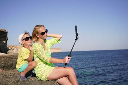 young blonde mom is relaxing with her little son on the sea on a Sunny clear day. A mother and child use a smartphone to make a video call on vacation.の写真素材
