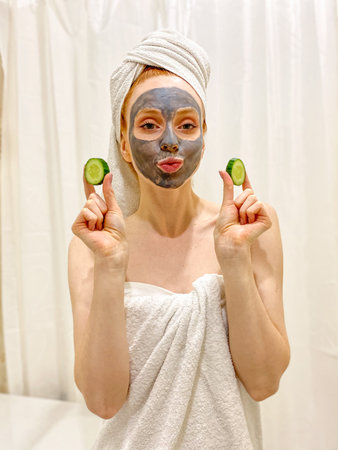 young woman with a towel on her head, a gray clay mask on her face, and a slice of cucumber in her...