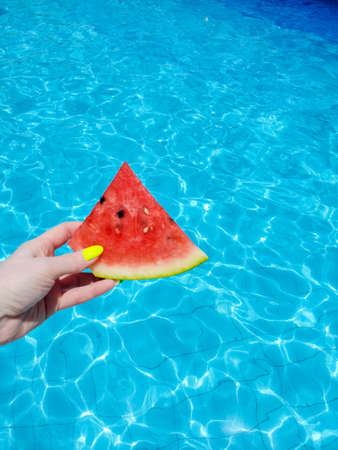 A slice of watermelon in a womans hand on the background of a blue poolの写真素材