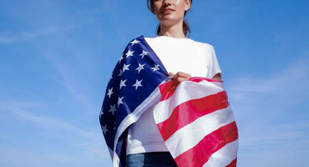 Portrait of a young woman with the national flag of the USA on her shoulders against the backdrop of a blue sunny sky. Young woman celebrating United States Independence Day.の写真素材