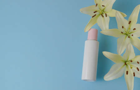 White empty cosmetic bottle with cream with pink cap, moisturizing lotion or shampoo framed with fresh lily flowers on a light blue background. Natural organic spa. Cosmetic concept. Top view.の写真素材