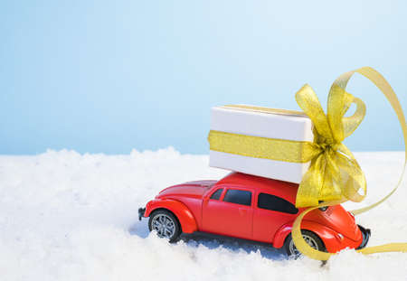 Volgograd, Russia - October 15, 2021: Small red retro toy car with a gift box with a gold ribbon and a Christmas tree on a blue background with white snow. Christmas, birthday, valentine's day, delivery concept.のeditorial素材