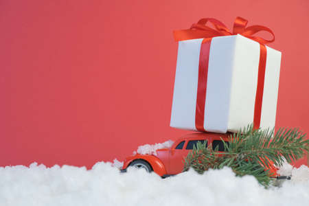 Volgograd, Russia - October 15, 2021: Red retro toy car delivering Christmas or New Year gifts and a Christmas tree tree on a festive red background and white snow.のeditorial素材