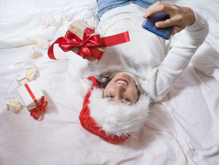Beautiful happy brunette woman makes a selfie by phone in a white sweater and a Christmas hat and...