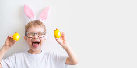 Happy child smiles in glasses in a white t-shirt in rabbit ears and with colorful yellow Easter eggs...