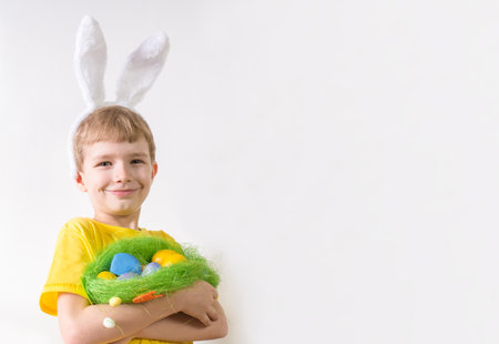 Happy child smiling in a yellow t-shirt in rabbit ears and with colorful eggs in a basket on a white...