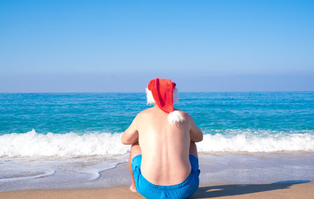 A man in a Santa Claus hat sits on the seashore, rear view. Christmas Santa Claus relaxing on the ocean tropical beach sand - New Year travel destinations, holidays in hot countries, travel discount concept.の写真素材