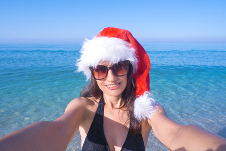 Christmas and holidays concept - Beautiful woman in Santas hat takes a selfie on the seashoreの写真素材