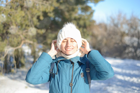 Portrait of a happy young brunette in winter clothes in a white knitted hat in the frost, in the sun...
