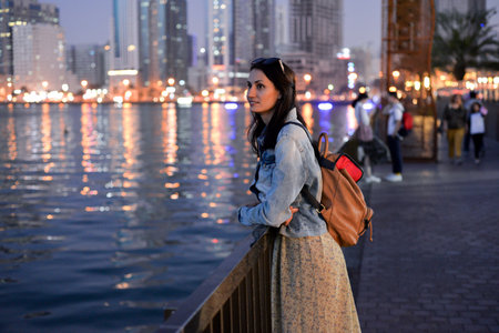 A tourist girl with a backpack on her shoulders enjoys a view of the modern skyscrapers of the Sharjah marina at nightの写真素材