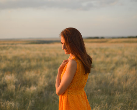 A pretty young girl with long brown hair in an orange dress in a field at sunset. The concept of freedom and natureの写真素材
