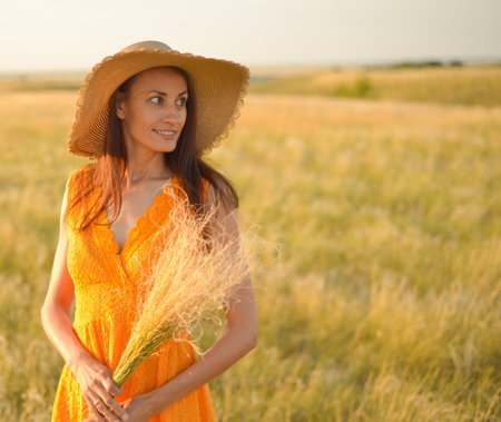 Young woman in an orange dress and a straw hat standing on a field in the rays of the setting sun.の写真素材