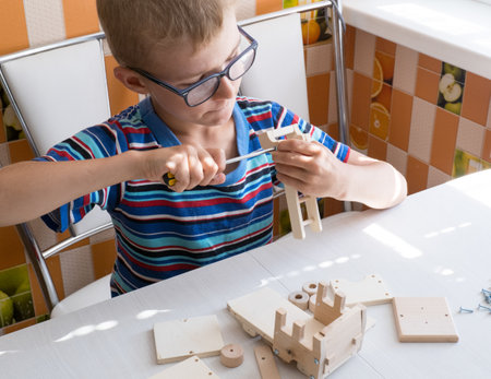 Portrait of a 7-8 year old boy with a screwdriver, carefully assembling a wooden car, sitting at the kitchen table. hands close up.の写真素材