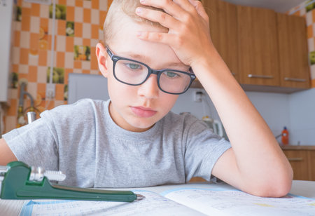 Tired sad little child boy sits at a table in the kitchen and writes homework. Boring lessons