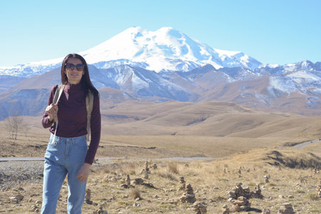 A female traveler with a backpack on her back admires the snow-covered Elbrus on a clear autumn day....
