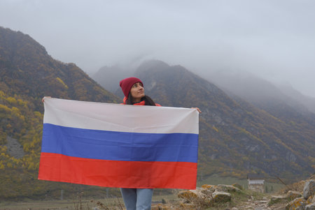Close-up of the Russian flag fluttering beautifully in the hands of a woman against the backdrop of the Caucasus Mountains on a cloudy foggy day. Russian Independence Day.の写真素材