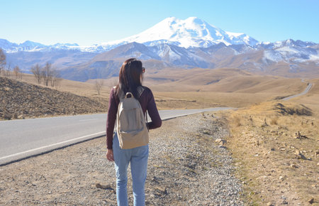 A female traveler with a backpack walks along a road in the mountains. View of Elbrus, North...
