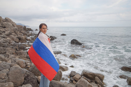 A brunette woman with a Russian flag on her shoulders stands on a rocky seashore. Independence day, patriotic holiday concept.の写真素材