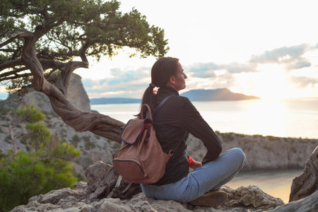 A young traveler with a backpack sits on the top of a mountain overlooking Cape Kapchik, Novy Svet...